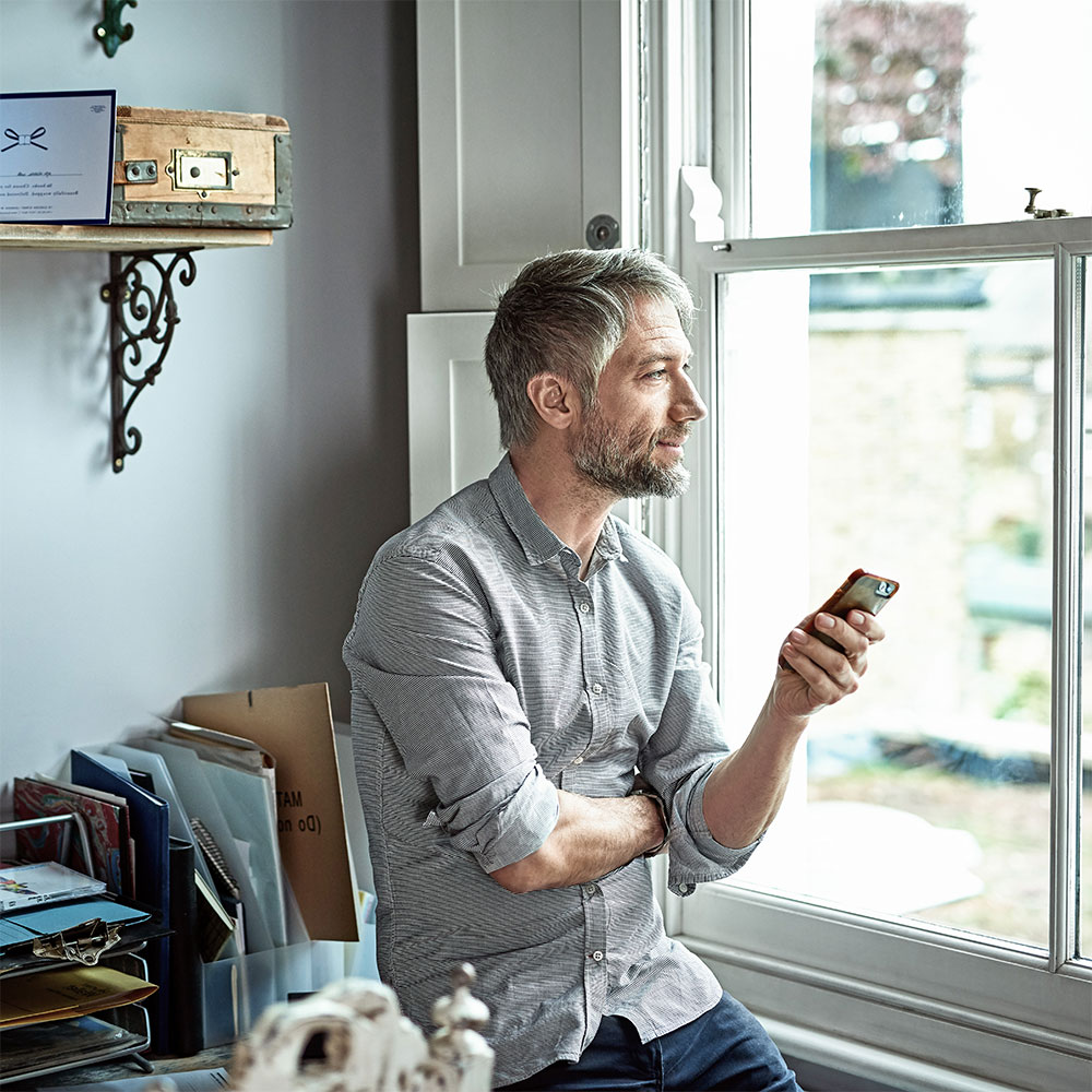 Mann schaut nachdenklich aus dem Fenster während er sein Smartphone in der Hand hält. Die Forschung zeigt: Es geht kein erhöhtes Krebsrisiko von Mobilfunkstrahlung aus. 