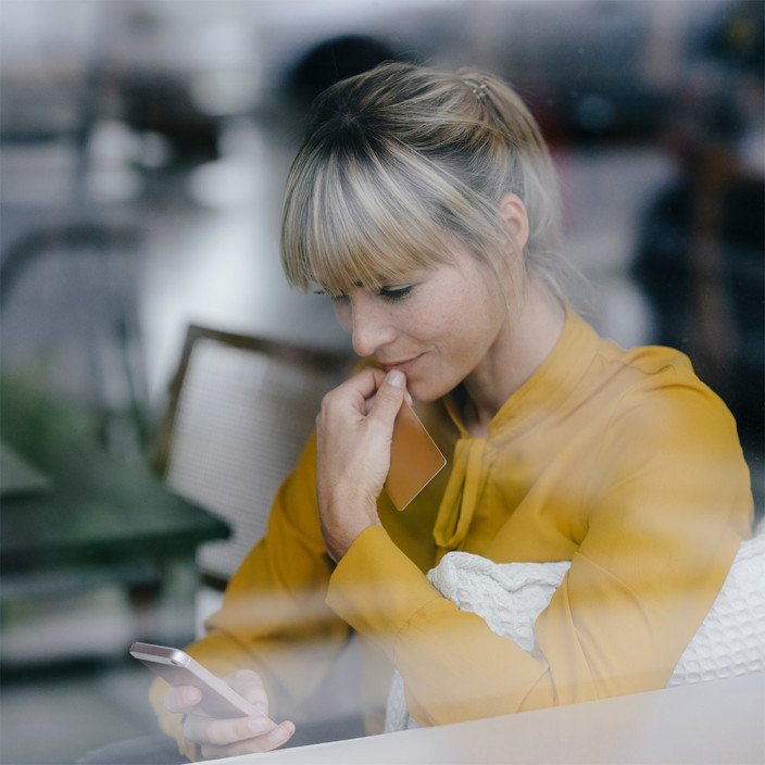 A woman in a yellow blouse behind a window pane is looking at her mobile device and is holding a card with her other hand. Many rules and laws ensure effective data protection and privacy for all citizens in the context of 5G mobile communications.