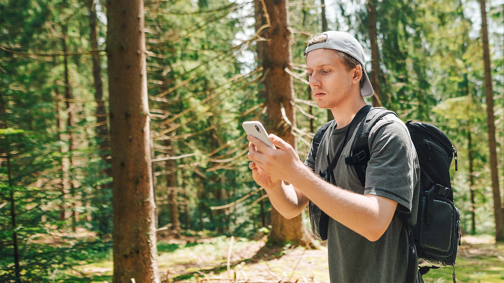 Mann mit Rucksack steht auf Waldlichtung und schaut auf sein Smartphone. Mitteilungen mit 5G schneller senden und empfangen – selbst mitten im Wald. 
