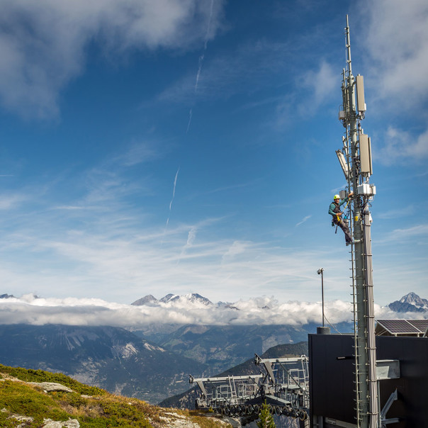 Mobilfunkmast ragt vor Bergkulisse in den blauen Himmel. Welche Bedingungen von Politik und Behörden geschaffen wurden und wie Betreiber das 5G-Netz aufbauen, erfahren Sie hier.