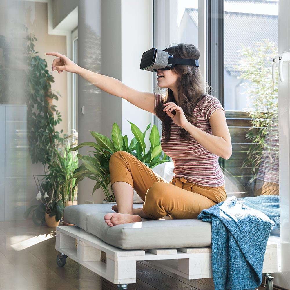 Frau sitzt auf Sofa und deutet mit dem Arm geradeaus während sie eine Virtual Reality Brille trägt. Mit dem 5G-Netz erkunden Nutzerinnen und Nutzer schnell und überall virtuelle Realitäten. 