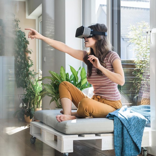 Frau sitzt auf Sofa und deutet mit dem Arm geradeaus während sie eine Virtual Reality Brille trägt. Mit dem 5G-Netz erkunden Nutzerinnen und Nutzer schnell und überall virtuelle Realitäten. 
