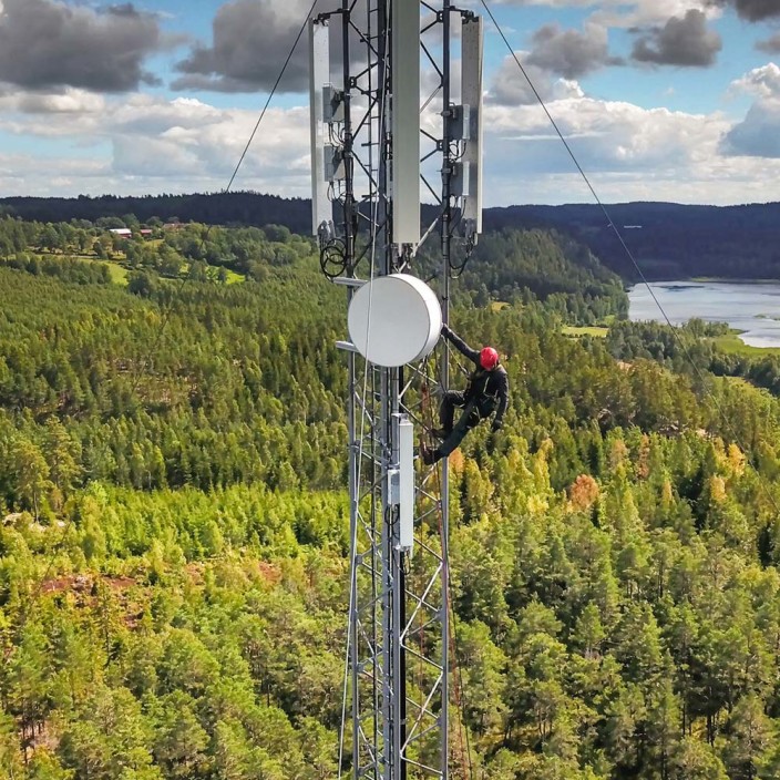 Ein Mobilfunk-Techniker klettert auf Sendemast vor einer Waldlandschaft. Ein 5G-Sendemast, der die umliegende Umgebung mit 5G Netz versorgt. 