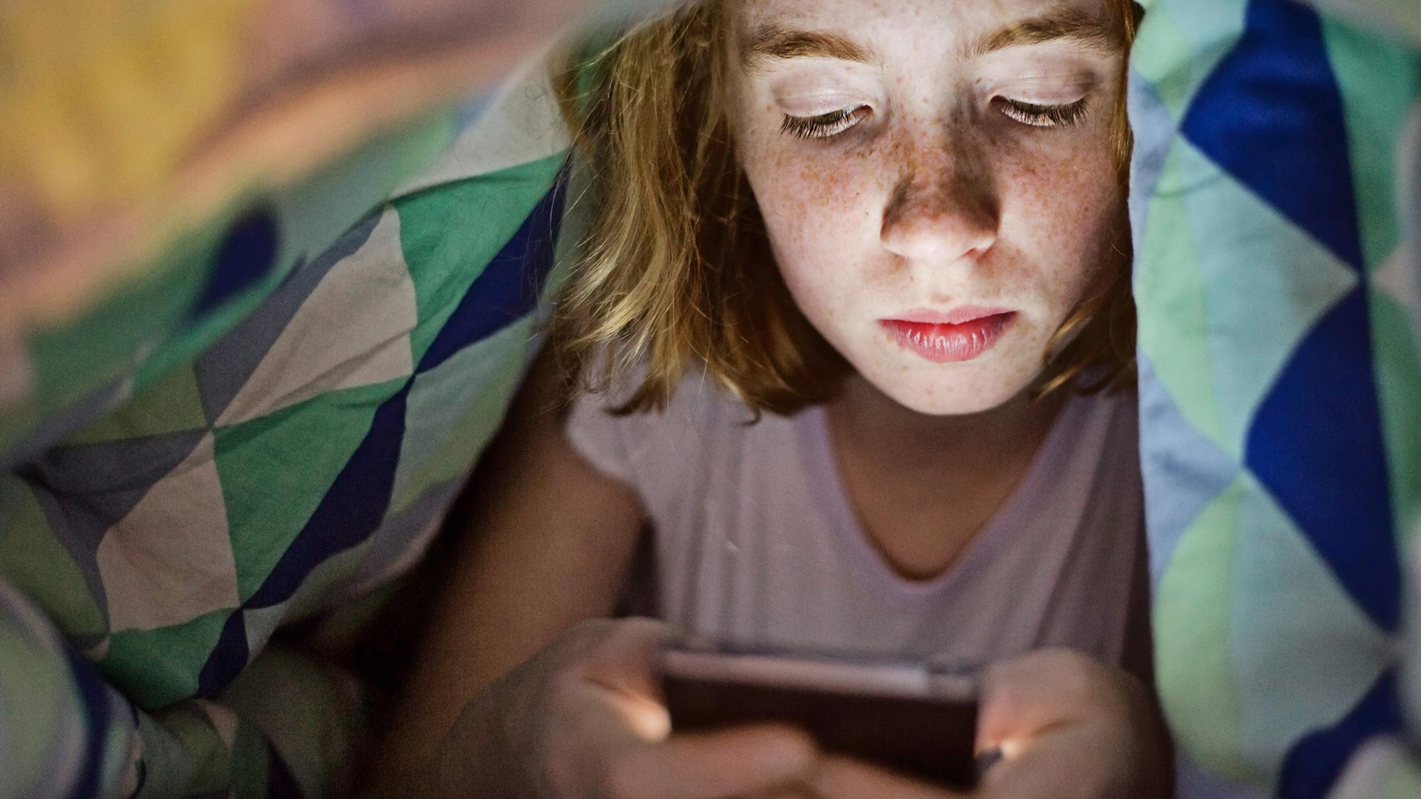 Mädchen mit Sommersprossen schaut auf ihr Smartphone unter der Bettdecke. Studien finden keine Belege für Schlaflosigkeit und Krankheiten durch elektromagnetische Felder. 