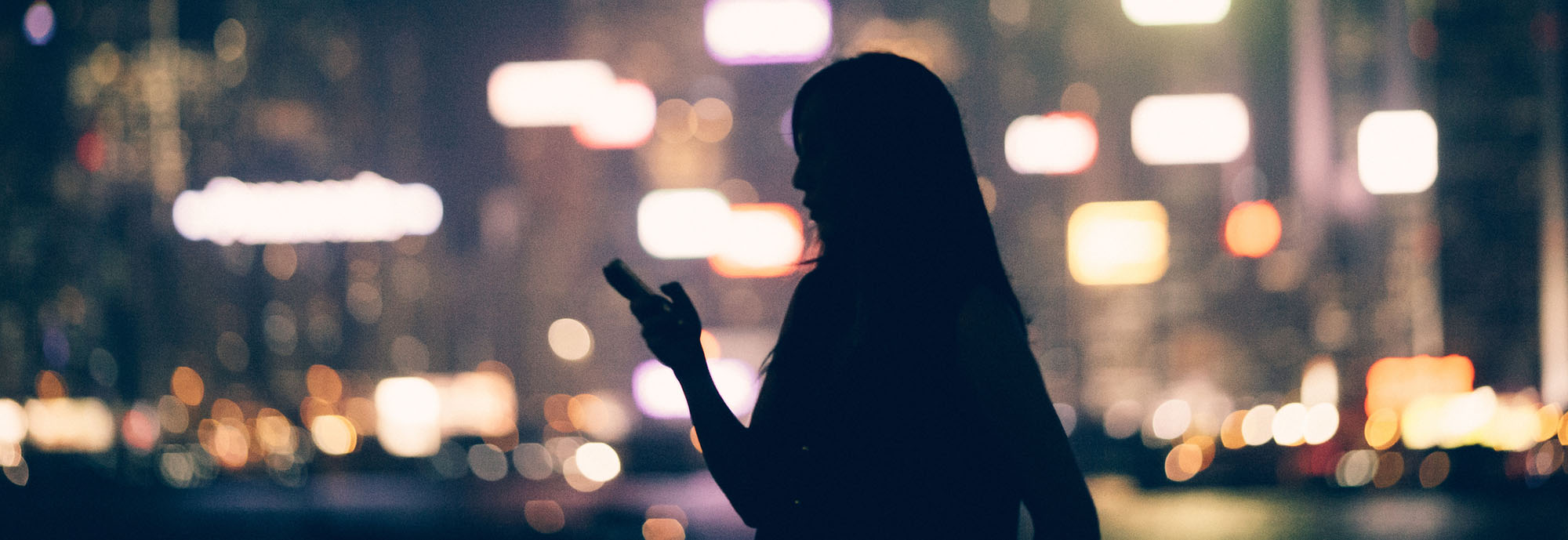 Die dunkle Silhouette einer Frau mit Mobiltelefon in der Hand vor den verschwommenen Lichtern der Stadt.