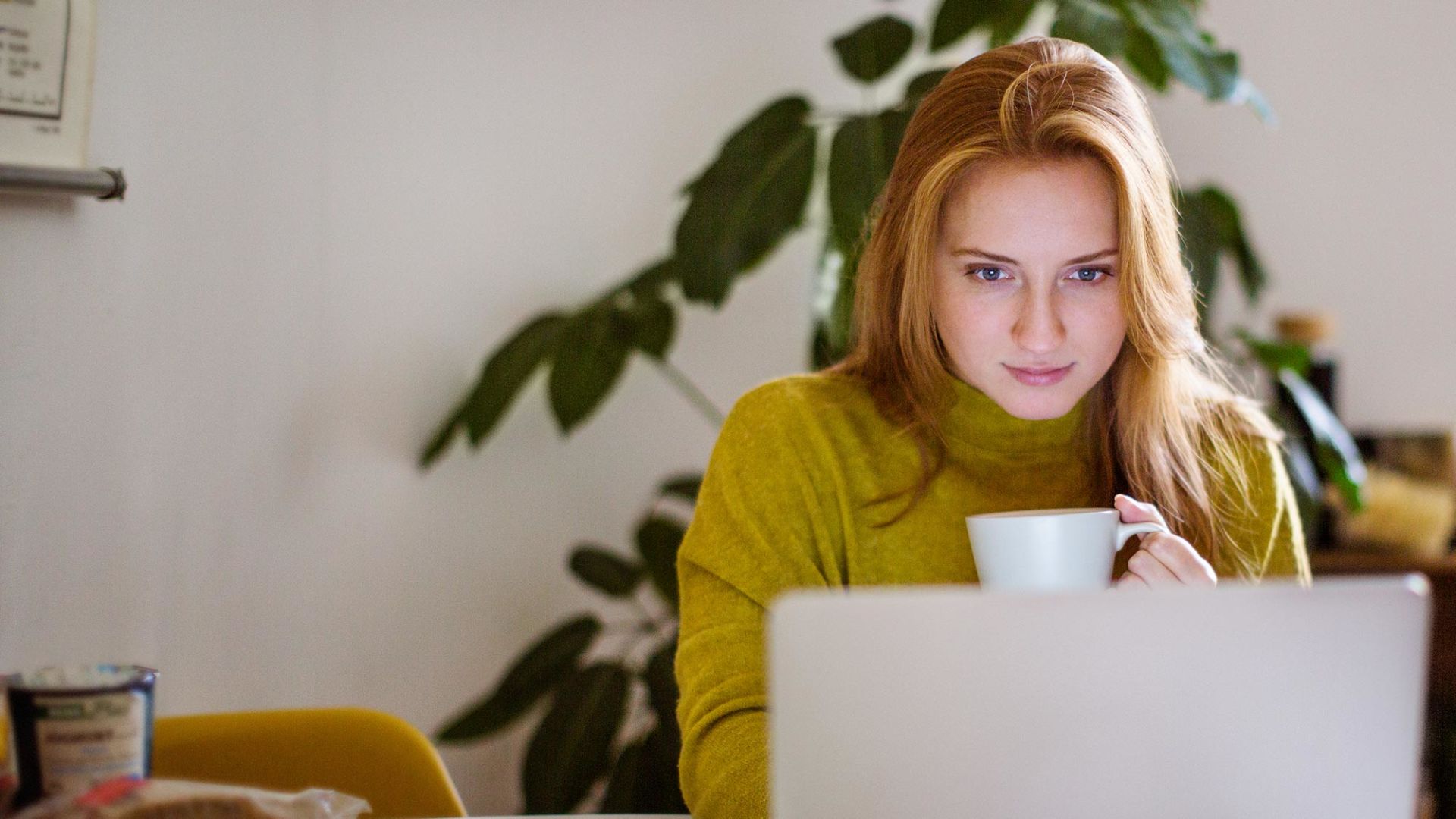 A woman is sitting at home in front of her laptop with a mug in her hand.