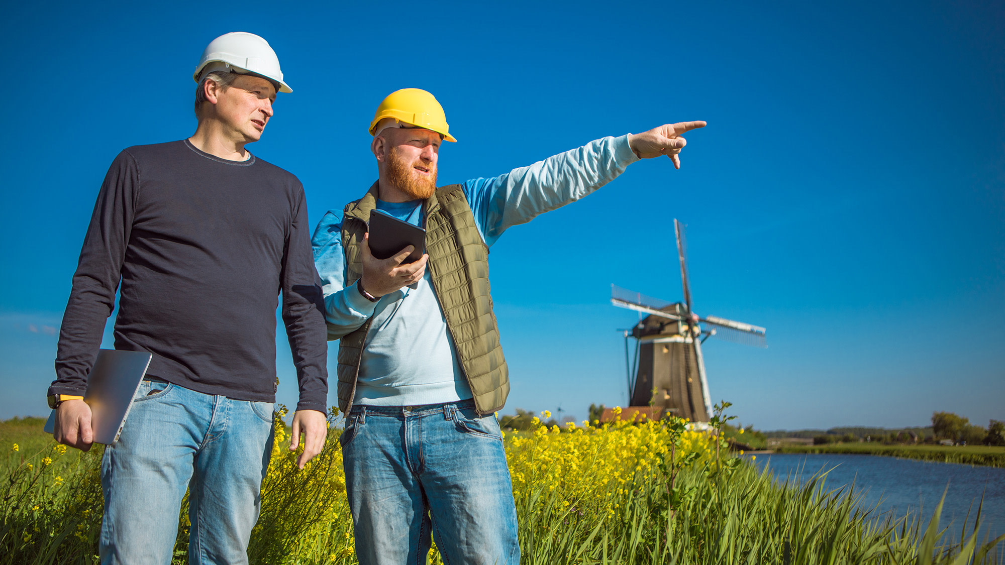 Zwei Männer mit Bauhelmen stehen vor einer Seenlandschaft mit einer Windmühle im Hintergrund.
