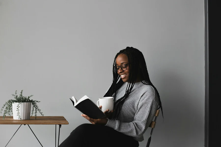A Black woman smiling, reading a book and holding a mug