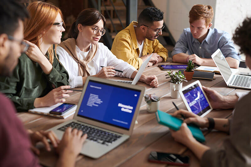 A group of glasses-wearing employees sit around a table with laptops during the workday.