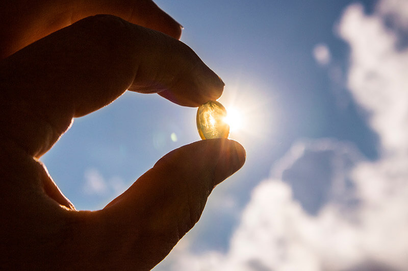 A hand holding a Vitamin D softgel pill up to the sunlight.
