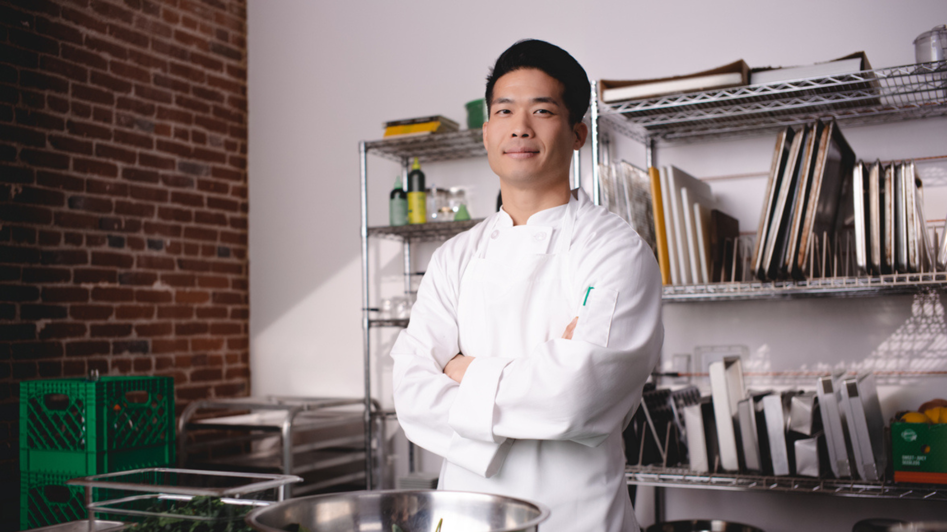 A chef smiles with their arms crossed, looking proud, standing over their prep station. 