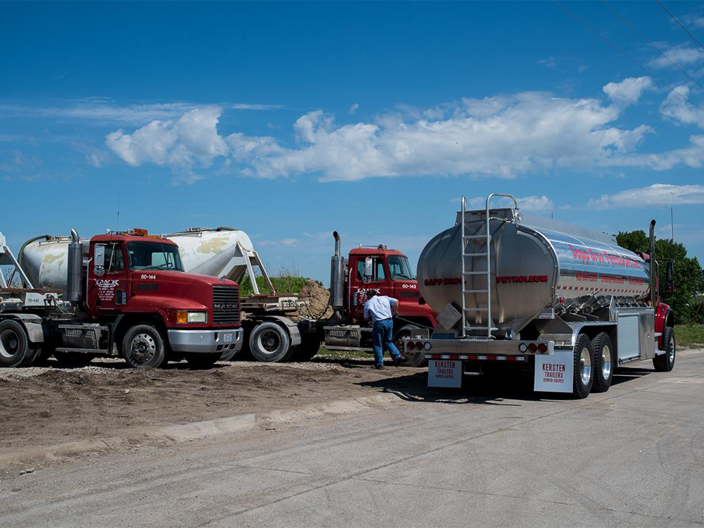 Sapp Bros. tanker trucks parked on a dirt lot under a blue sky, with a worker standing near one vehicle.