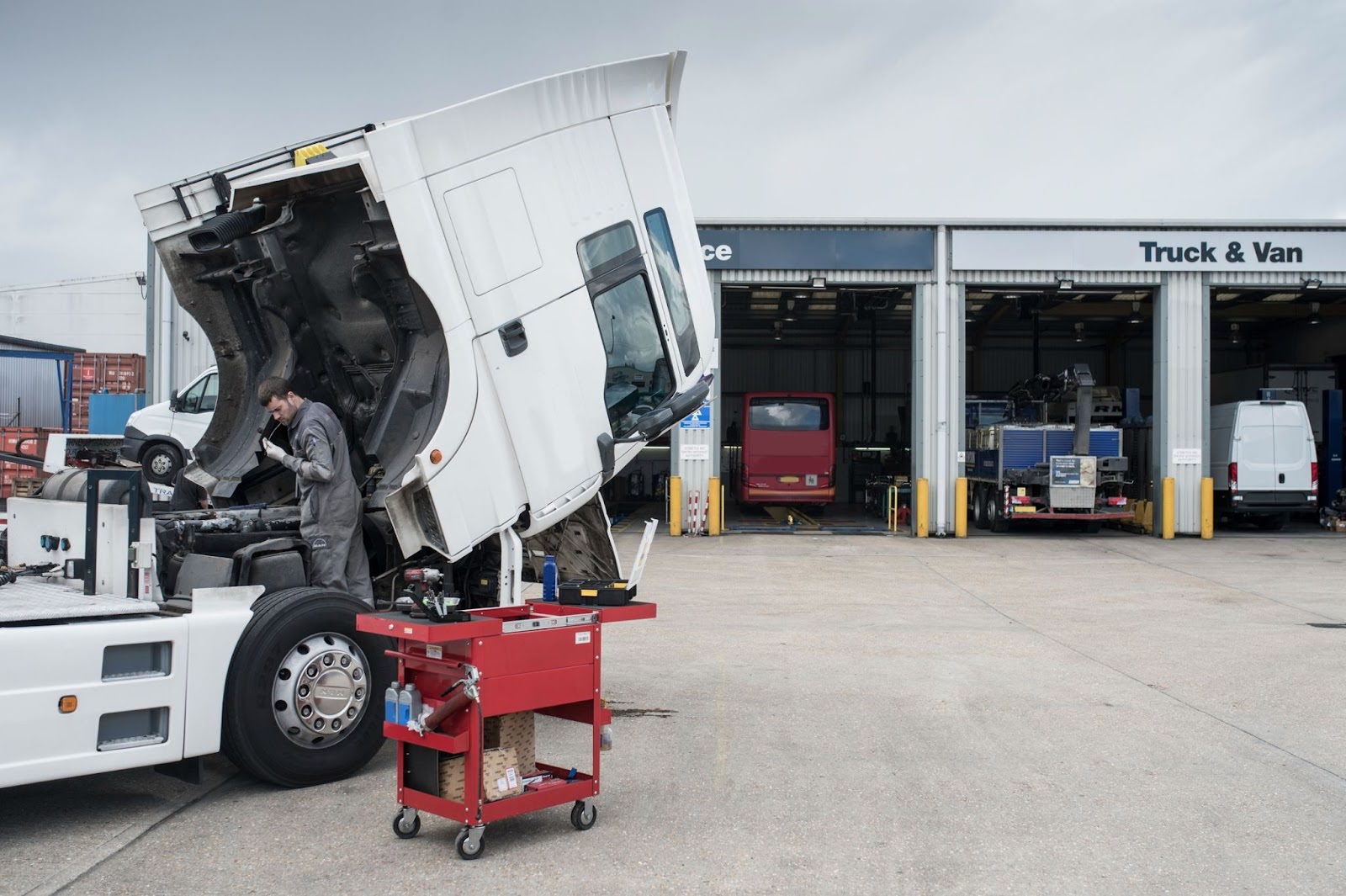 Mechanic working on white semi truck with tilted cab at a truck service center with repair bays visible in background.