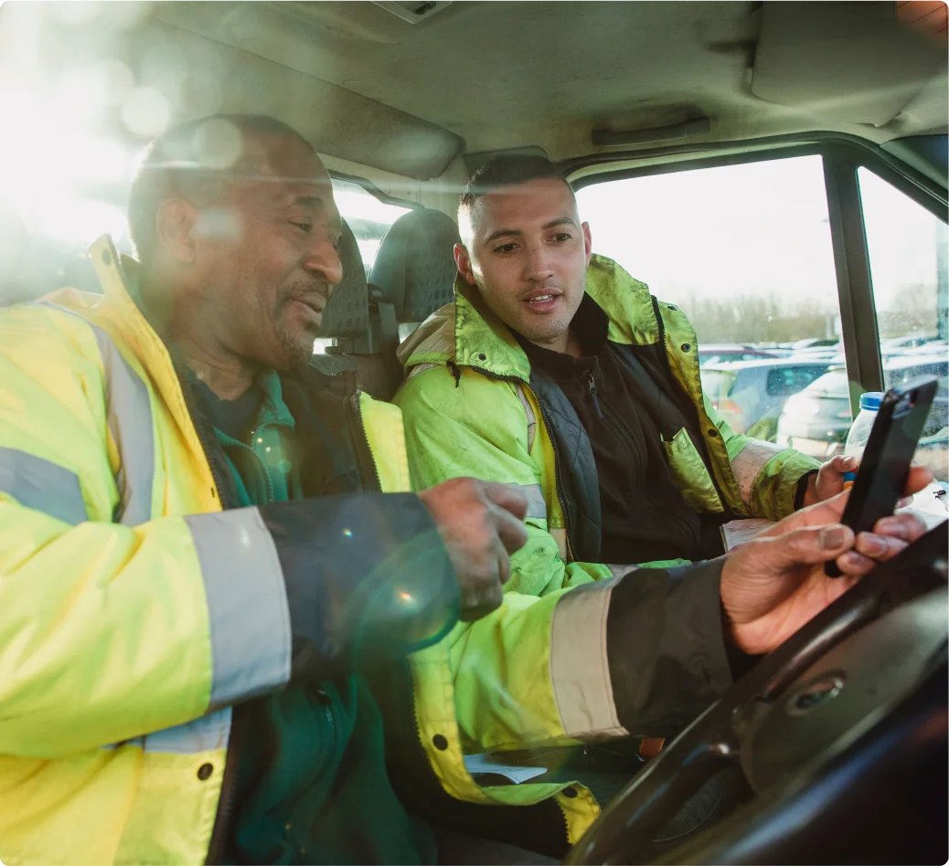 Dos trabajadores con chaleco de de alta visibilidad viendo un dispositivo tipo tableta dentro de un vehículo.