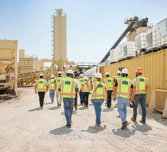 Construction workers in bright yellow safety vests and hard hats walking through an industrial cement plant site.