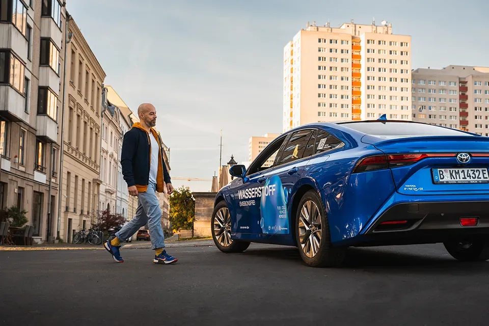 Mann geht auf einen blauen Toyota Mirai Wasserstoff-Auto in einer Stadtstraße zu, Gebäude im Hintergrund.
