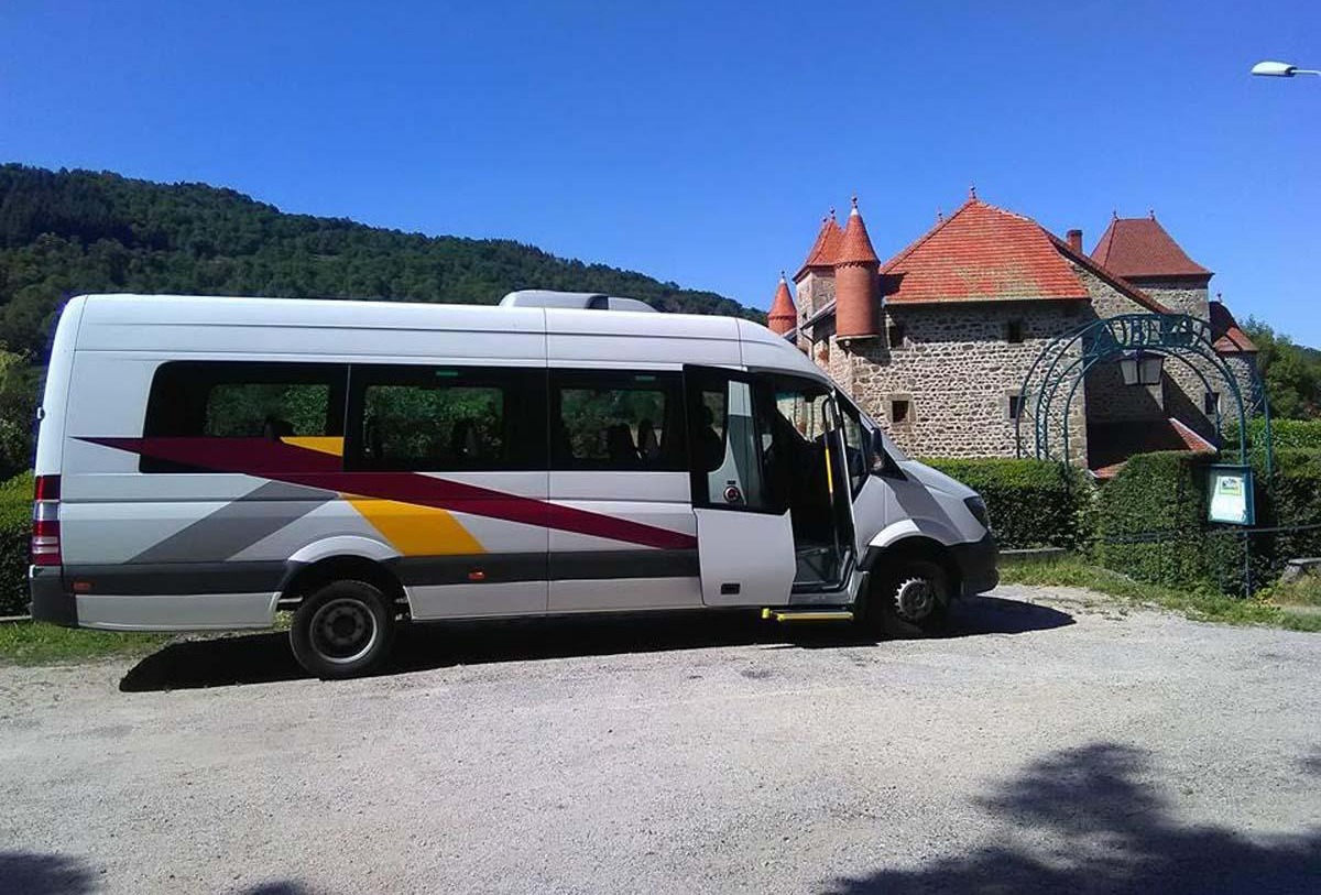 Minibus blanc avec décorations colorées stationné devant un château médiéval en pierre avec toits rouges sous un ciel bleu.
