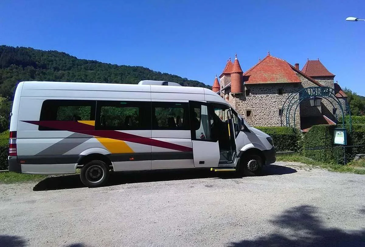 Minibus blanc avec décorations colorées stationné devant un château médiéval en pierre avec toits rouges sous un ciel bleu.