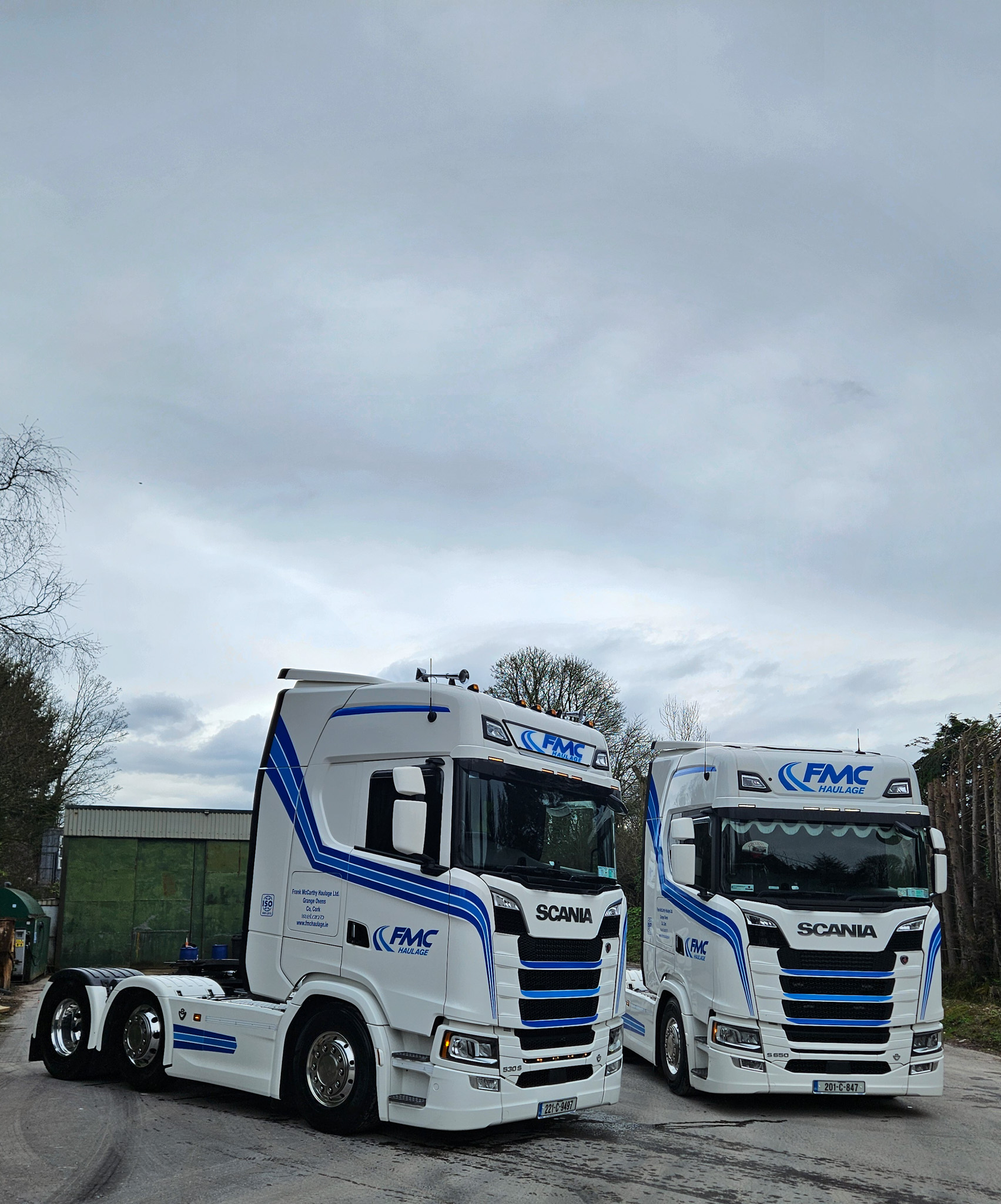 Two white Scania trucks with blue FMC Haulage livery parked side by side under a cloudy sky.