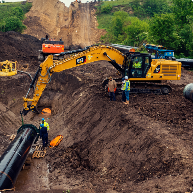 Yellow excavator and workers installing large black pipeline in a hillside trench at a construction site.