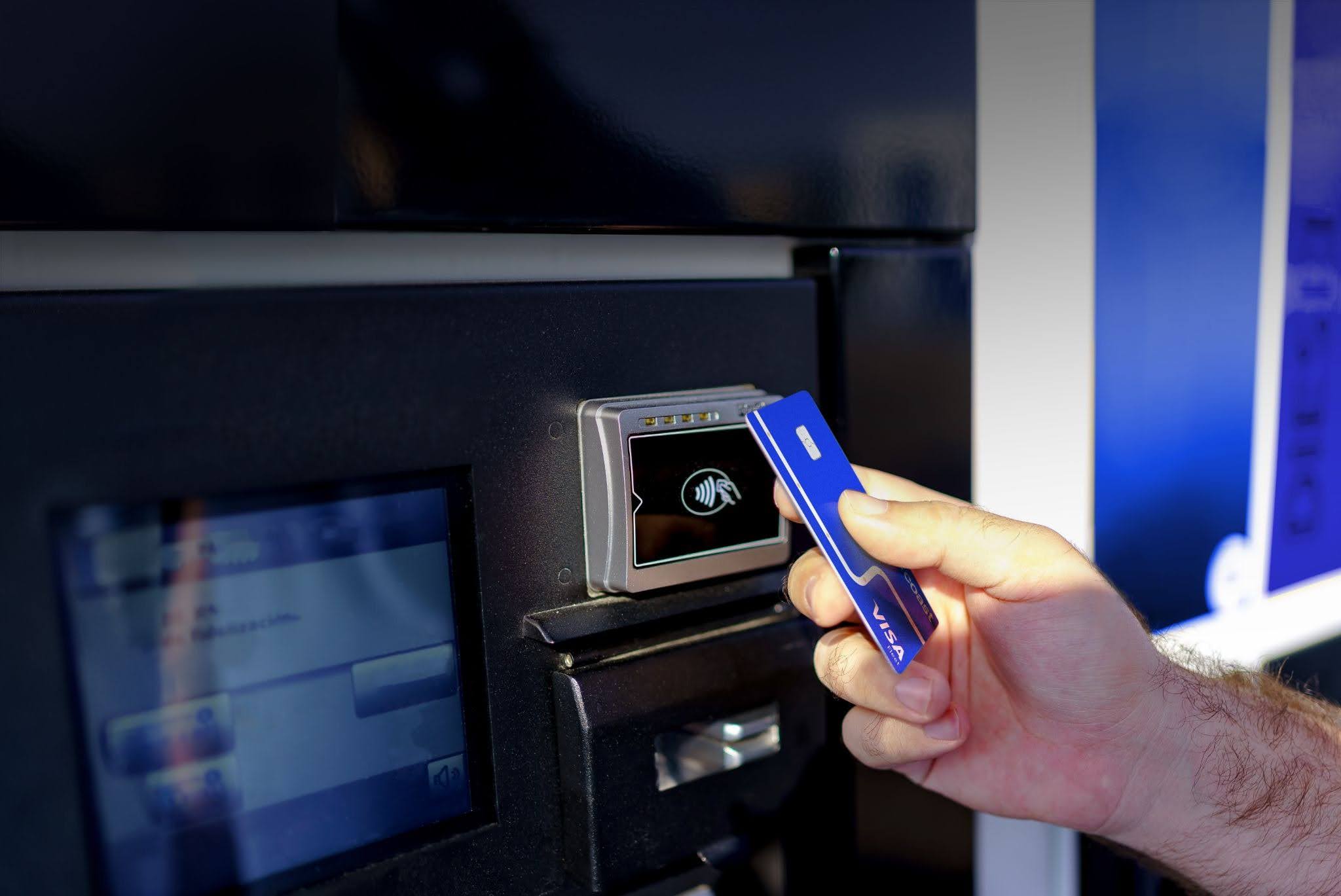 A hand tapping a blue Visa card on a contactless payment reader at a self-service kiosk or ATM machine.