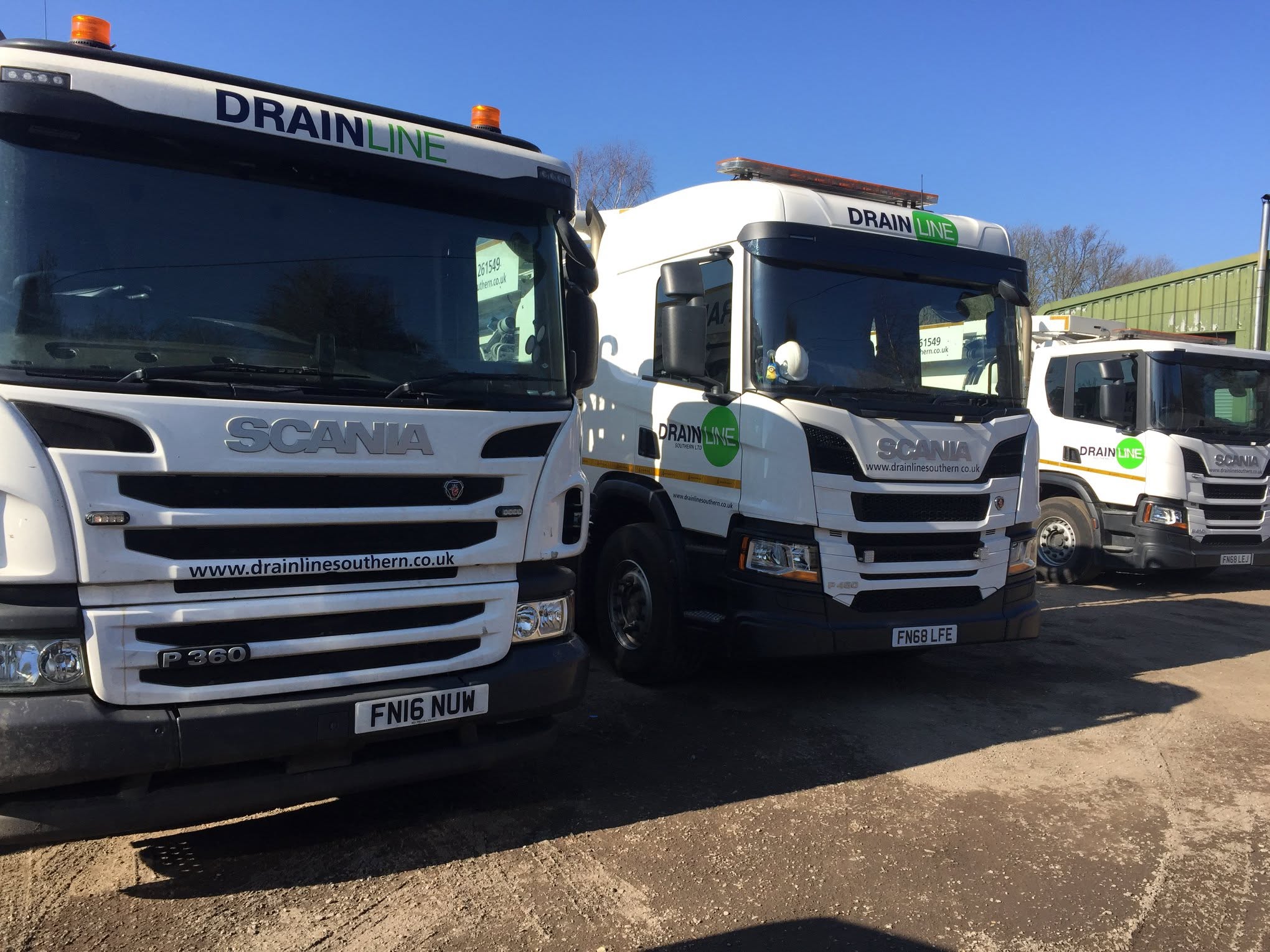 Three white Scania trucks branded with Drainline Southern logos parked in a yard under a clear blue sky.