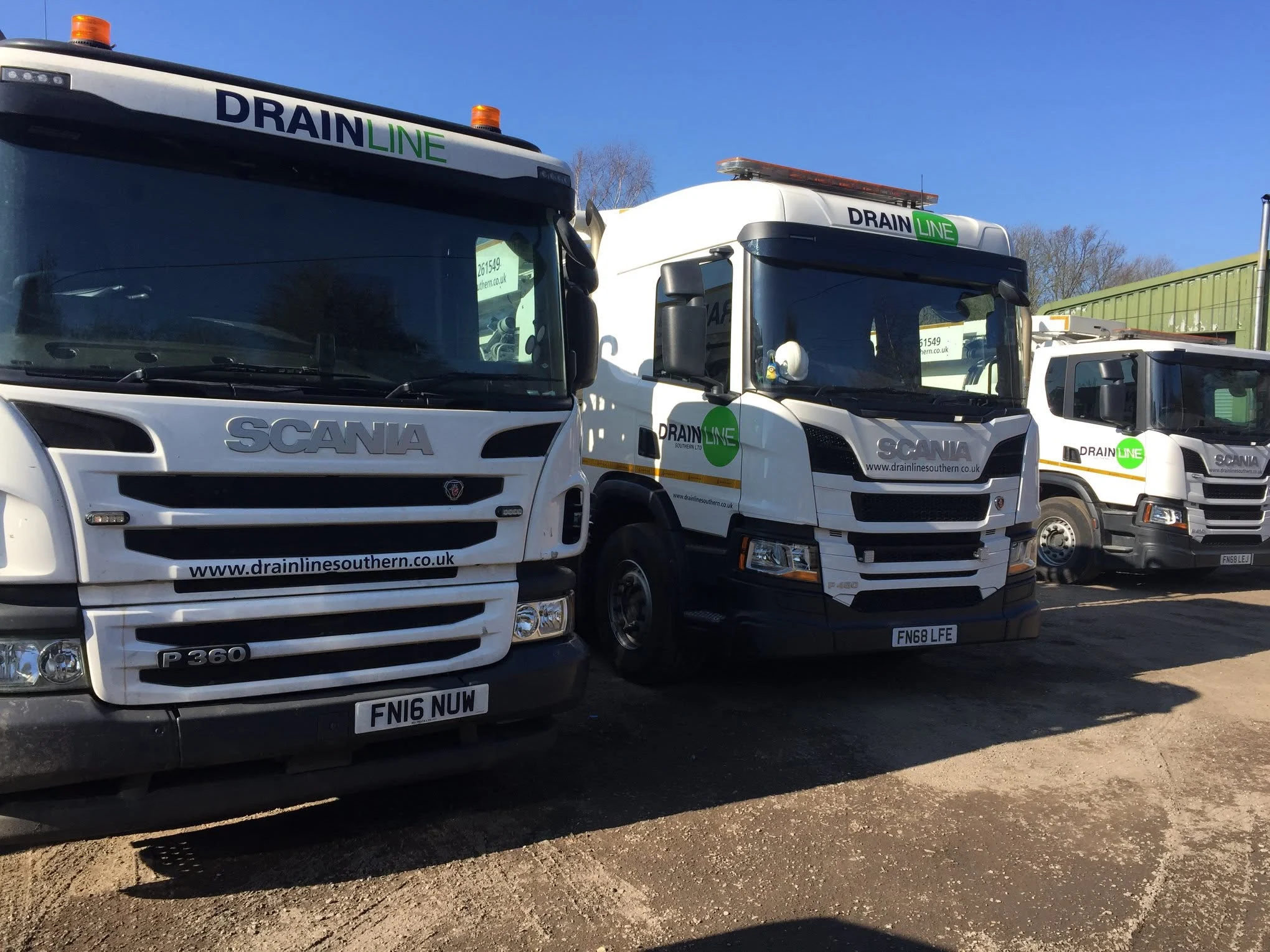 Three white Scania trucks branded with Drainline Southern logos parked in a yard under a clear blue sky.