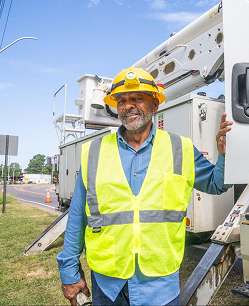 Construction worker in yellow hard hat and safety vest standing next to a utility truck on a roadside.