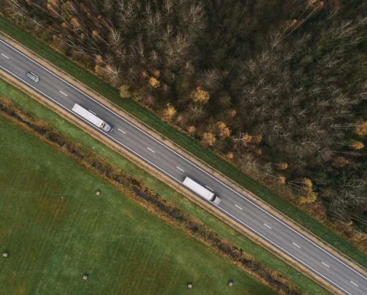 Overhead view of two trucks driving down a highway with woods on one side and an open field on the other