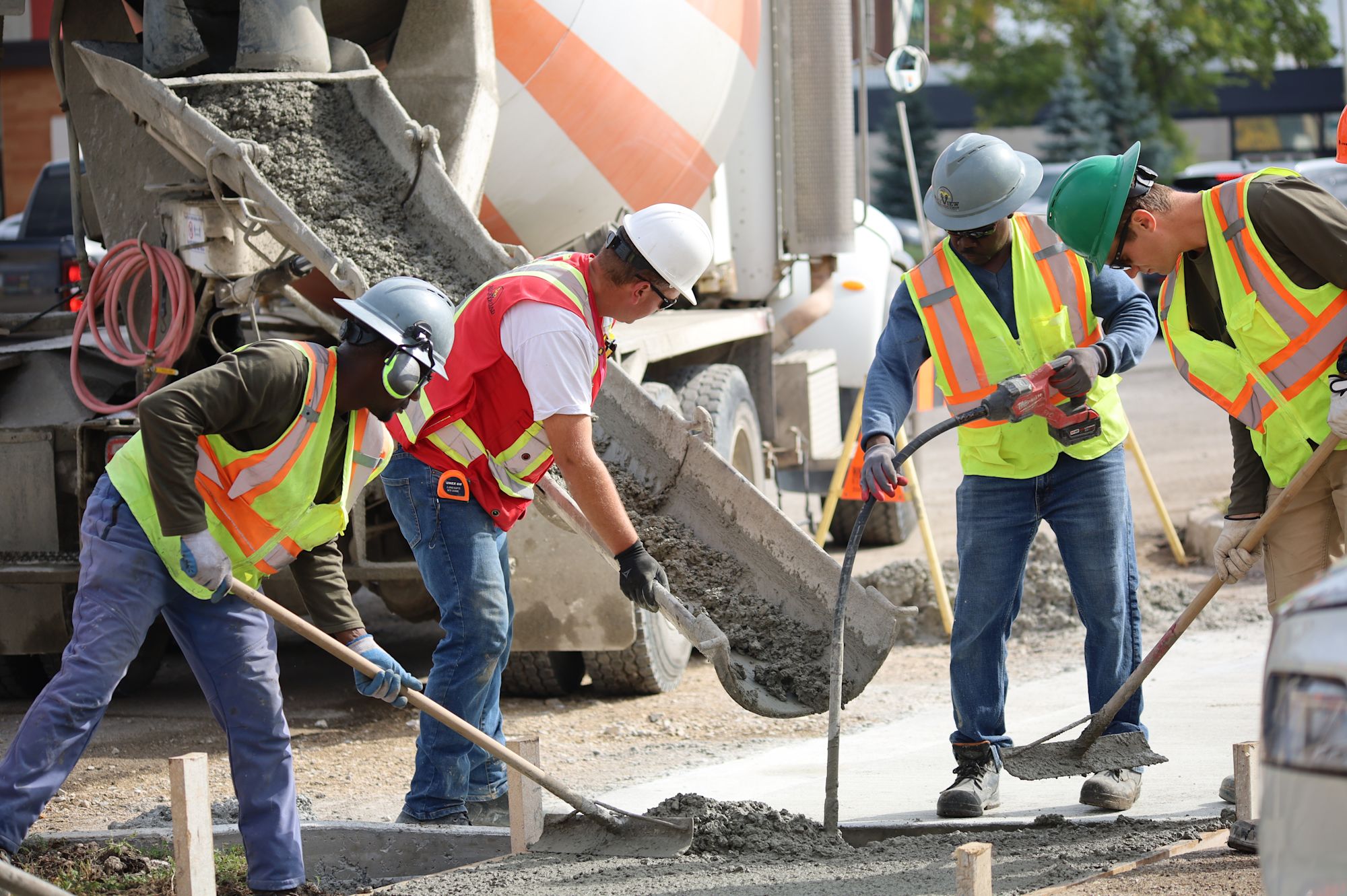 Construction workers in safety vests and hard hats pouring and spreading concrete from a cement mixer truck at a job site.