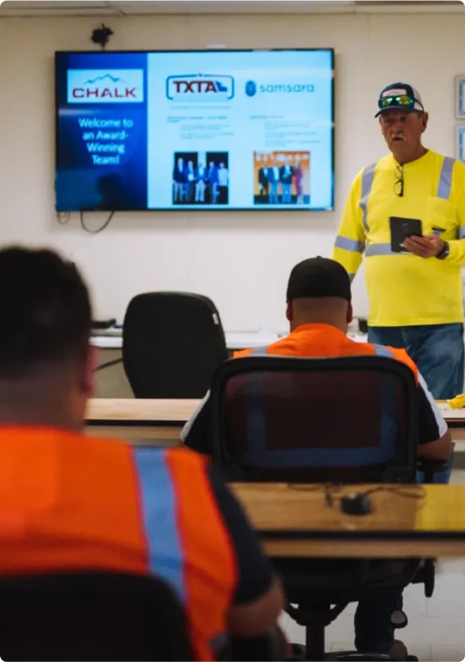 Trabajador con chaleco de seguridad amarillo presenta a sus colegas con chalecos naranjas, con pantalla que muestra logotipos