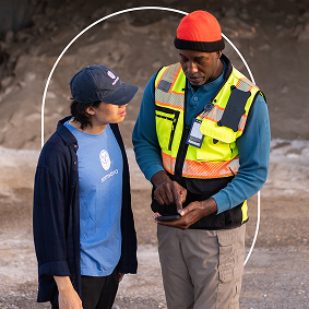Two people conversing outdoors; one wearing safety vest and orange hat, the other in blue shirt and cap.