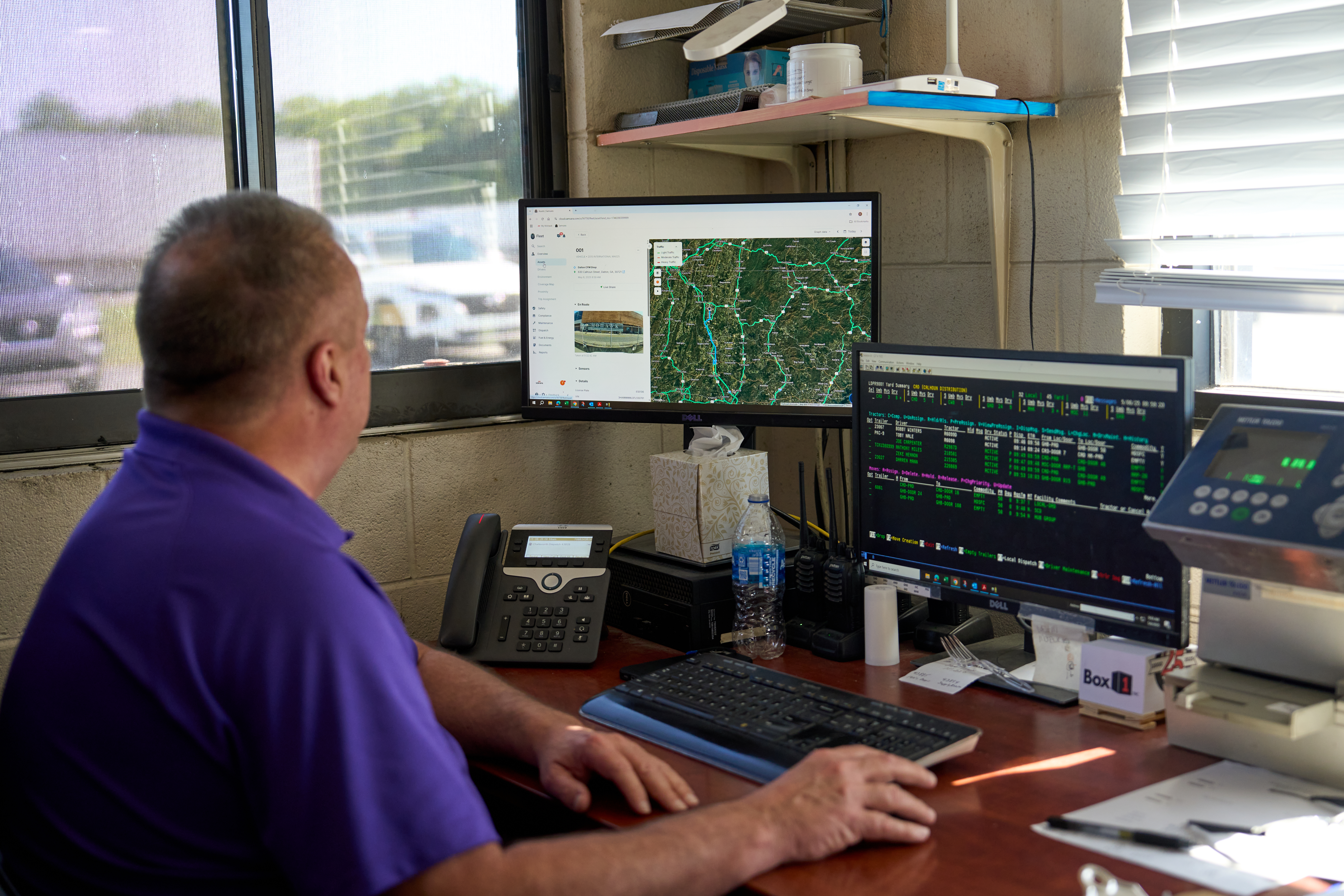 Person in purple shirt monitoring multiple computer screens showing maps and data in a dispatch or control room setting.