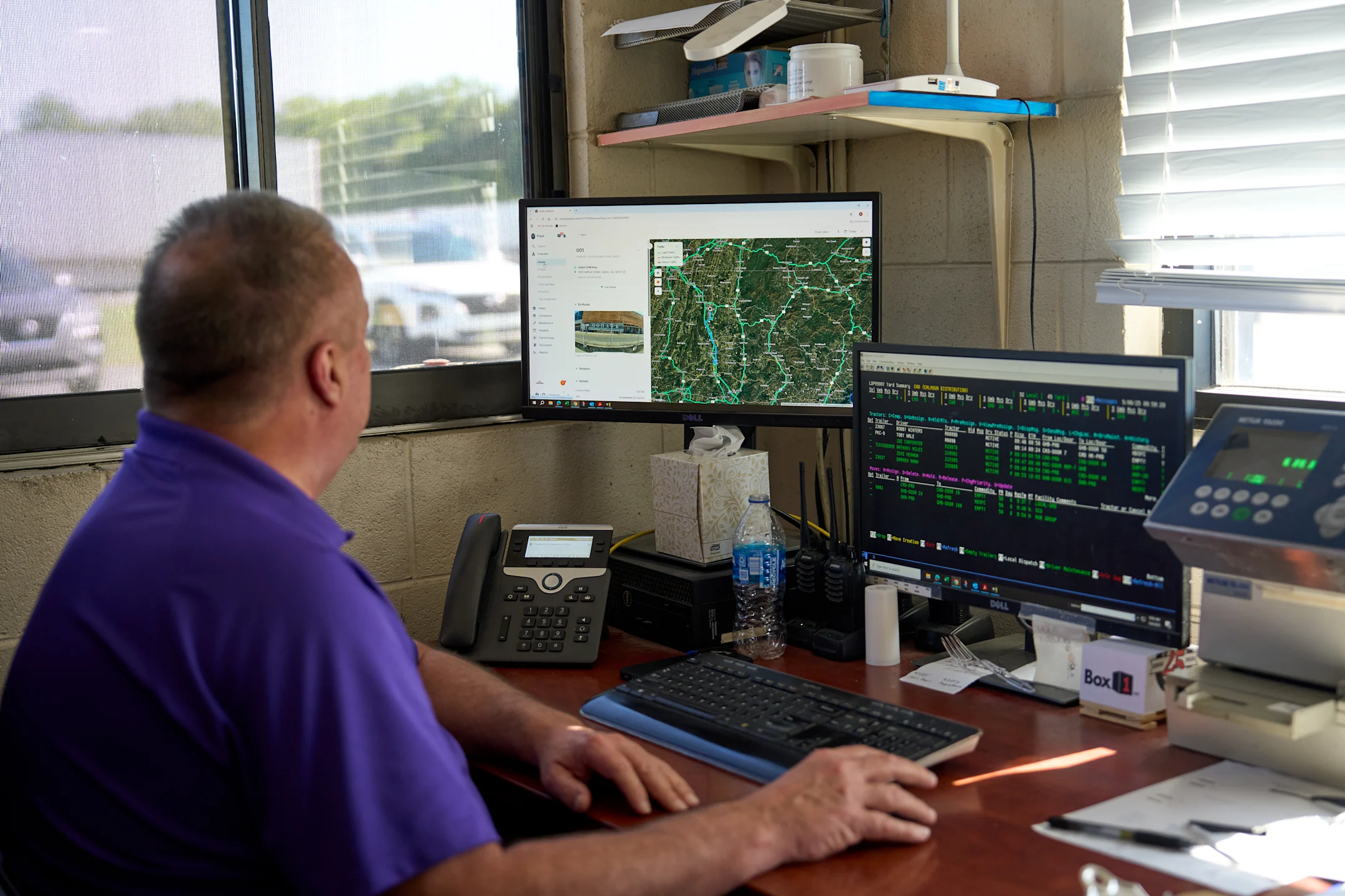 Person in purple shirt monitoring multiple computer screens showing maps and data in a dispatch or control room setting.