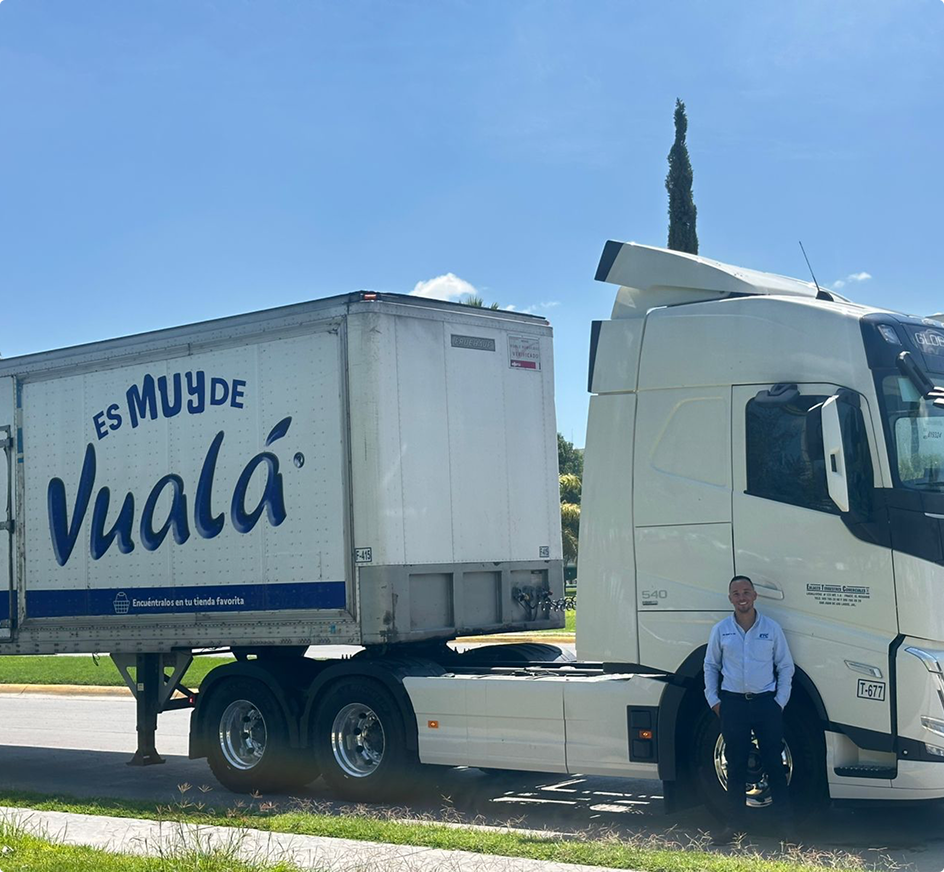 Camión de carga blanco con remolque que dice "ES MUY DE Vualá" estacionado bajo cielo azul con una persona junto a él.