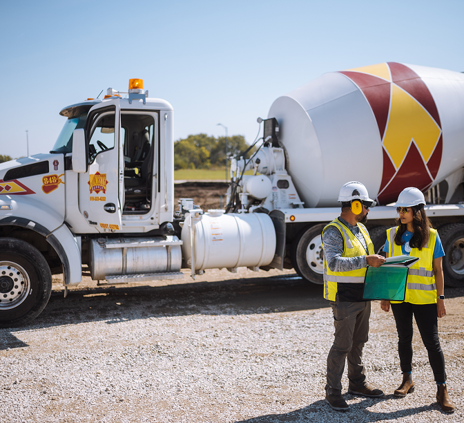 Two construction workers in safety gear reviewing documents beside a cement mixer truck at a gravel worksite.