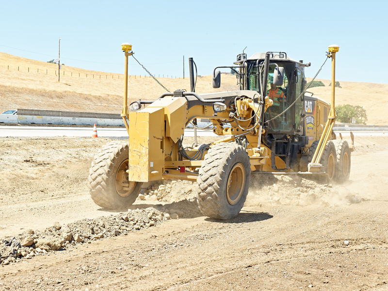A yellow CAT motor grader operates on a dusty construction site, grading dry dirt near a highway on a sunny day.