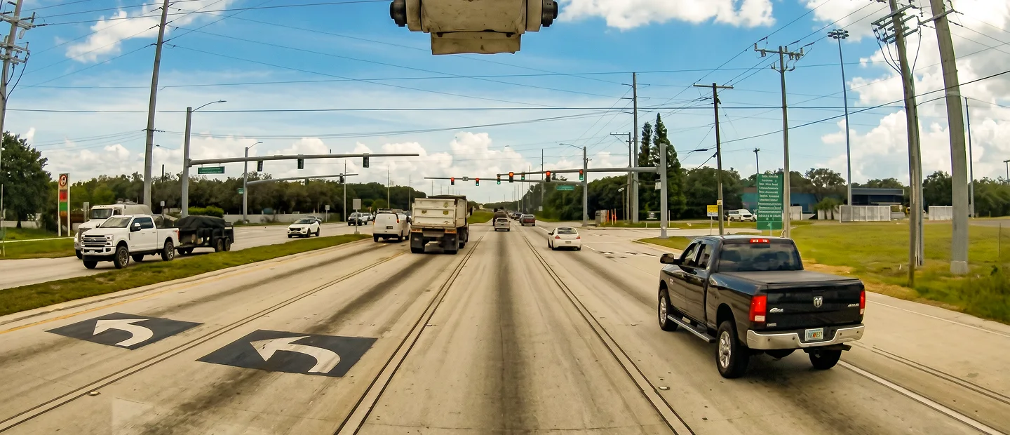 A busy multi-lane road with trucks and cars under a blue sky, traffic signals overhead, and green signs along the roadside.