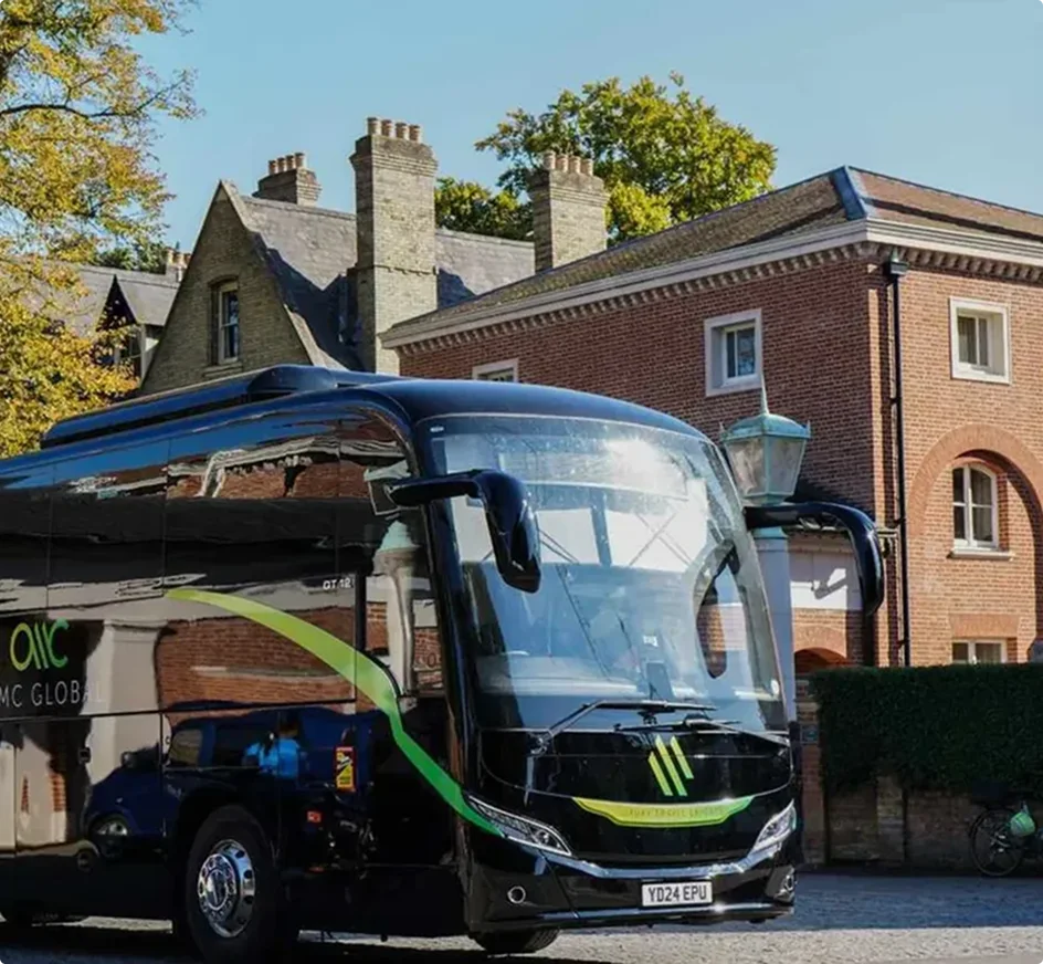 Black luxury coach with green accents parked in front of a brick building with multiple chimneys on a sunny day.