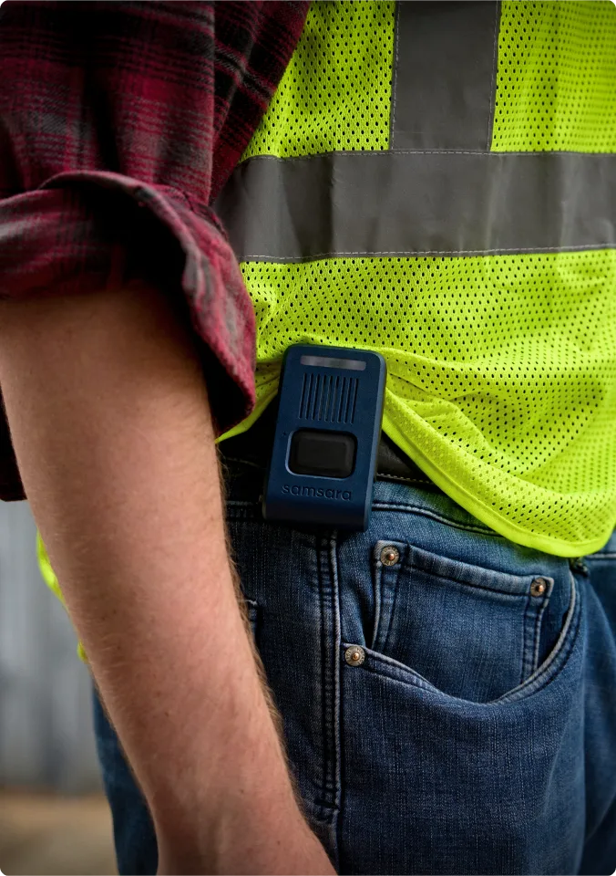 Person wearing red plaid shirt and high-visibility safety vest with blue Samsara device clipped to jeans pocket.