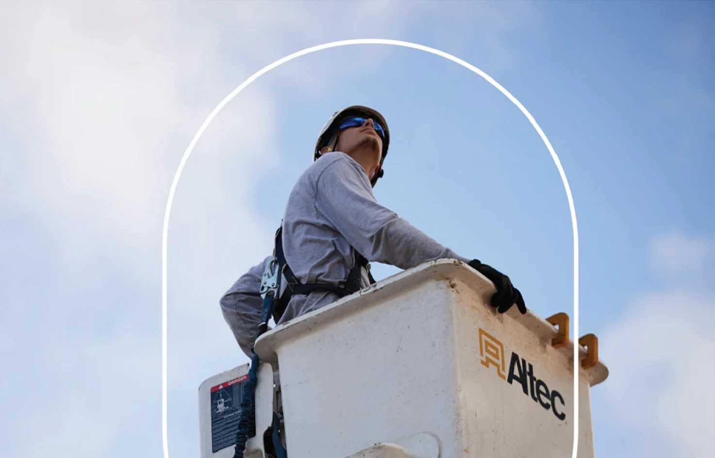 A construction worker wearing protective gear stands in an elevated bucket truck against a blue sky