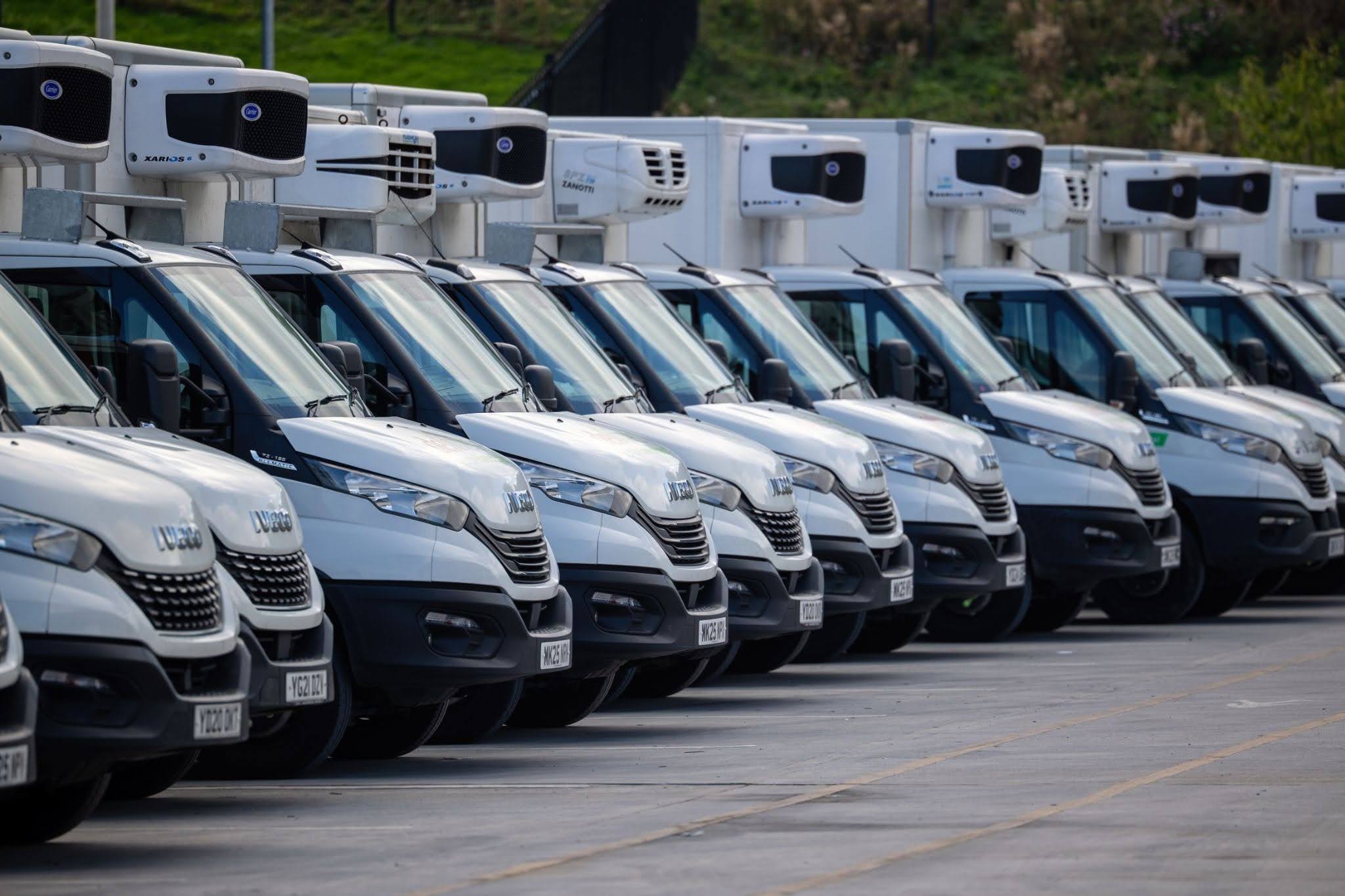 A fleet of white Iveco refrigerated vans with rooftop cooling units parked in rows in a large car park.