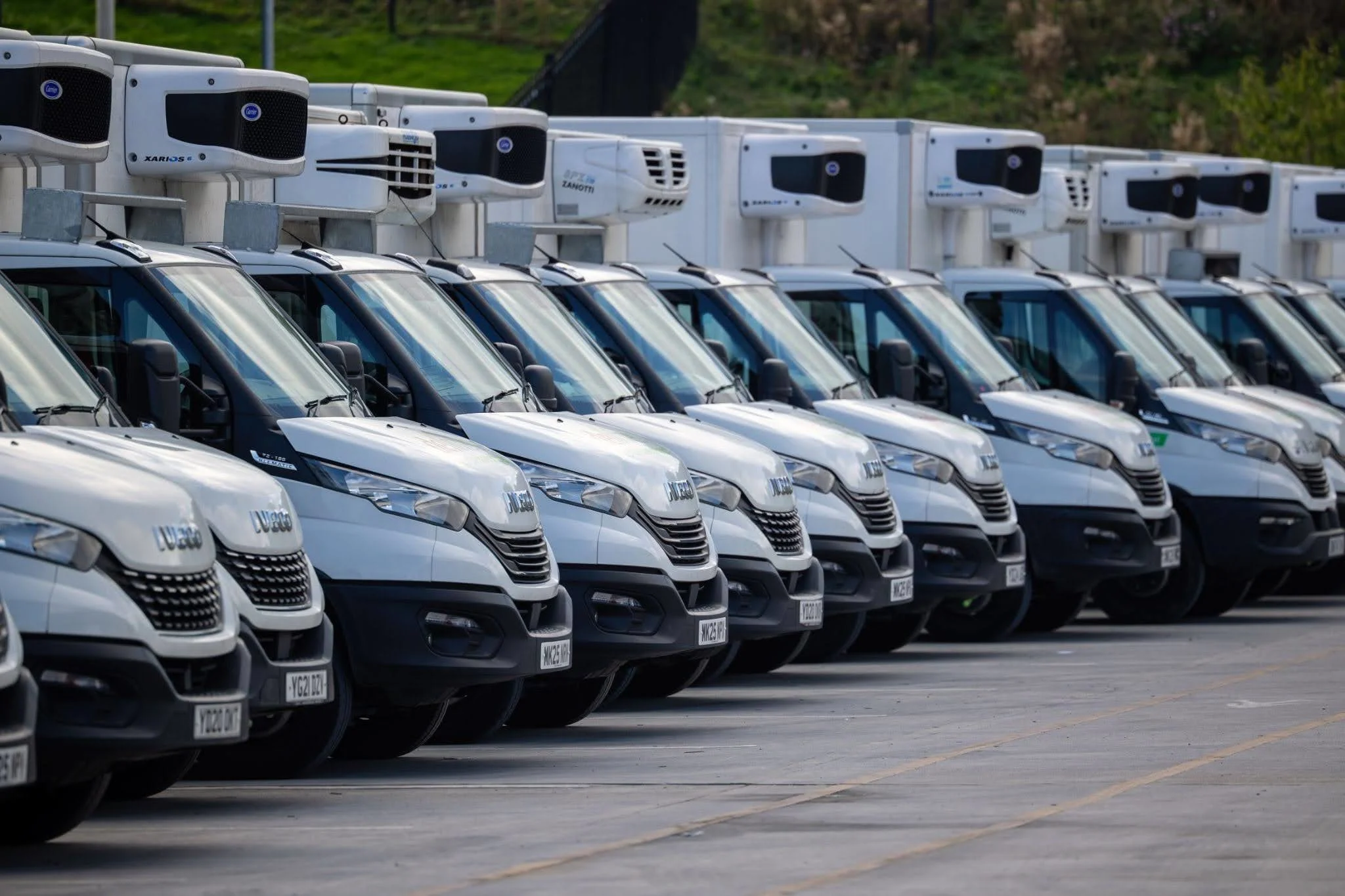 A fleet of white Iveco refrigerated vans with rooftop cooling units parked in rows in a large car park.