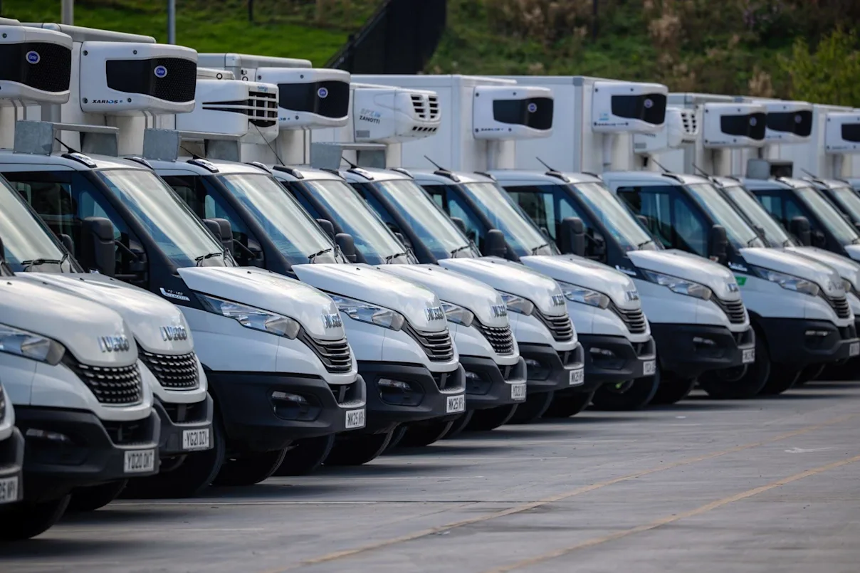 A fleet of white Iveco refrigerated vans with rooftop cooling units parked in rows in a large car park.