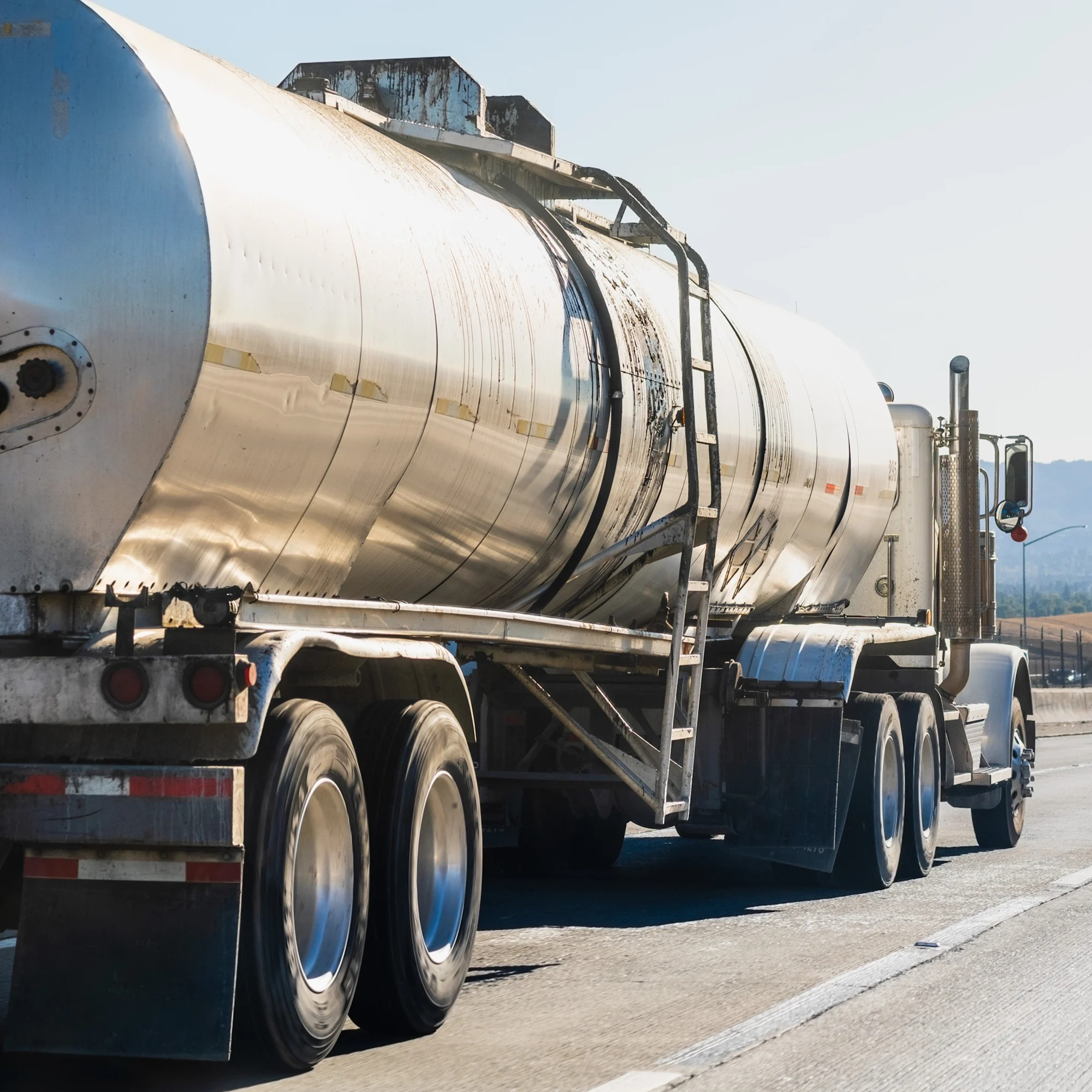 Camión cisterna plateado con número 295 transportando material inflamable en carretera con montañas al fondo bajo cielo azul.