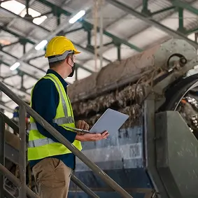 Worker in yellow hard hat and safety vest inspecting industrial equipment with clipboard in a factory.