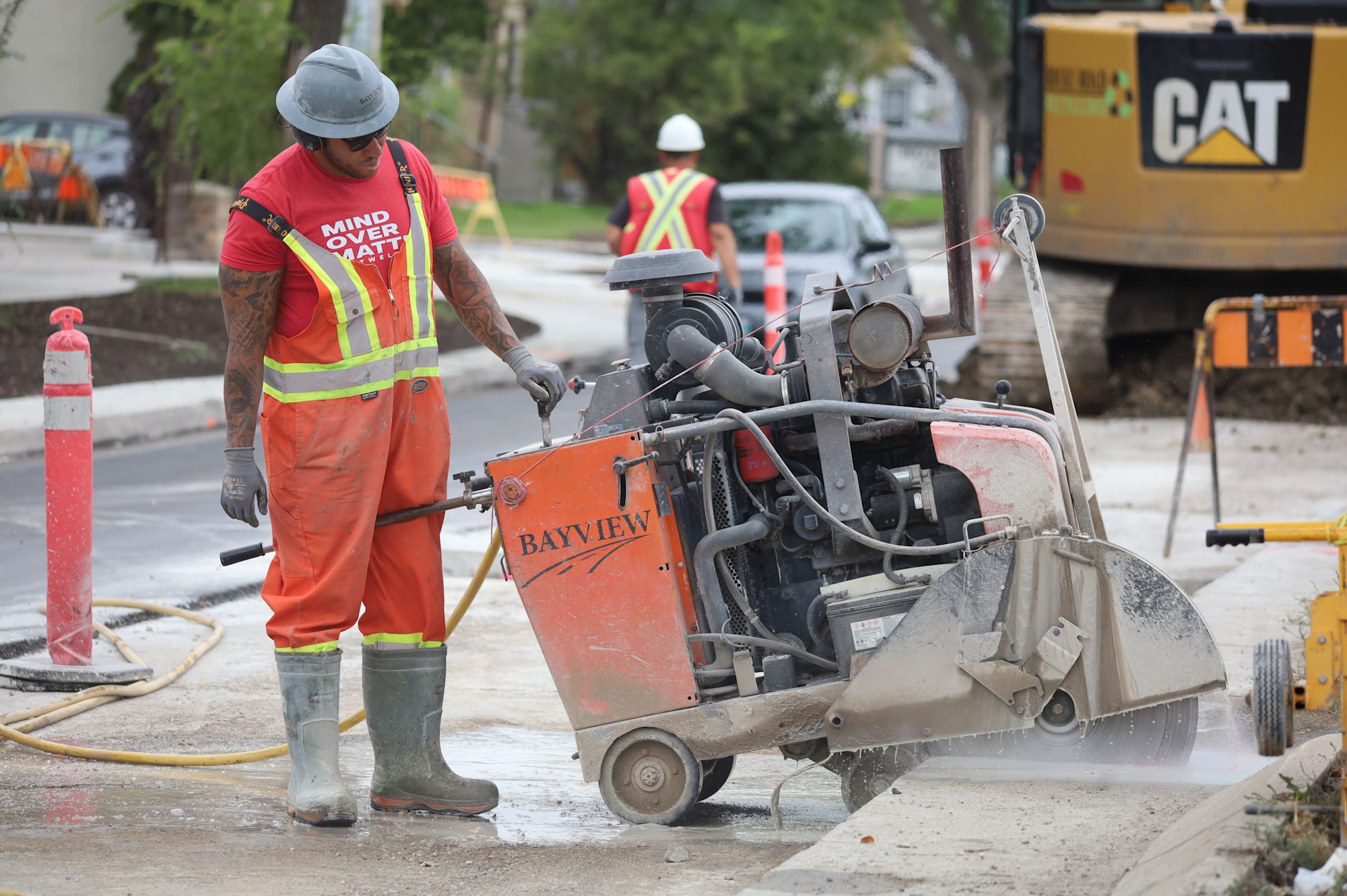Construction worker in bright safety gear operating a concrete saw on a street with CAT equipment nearby.