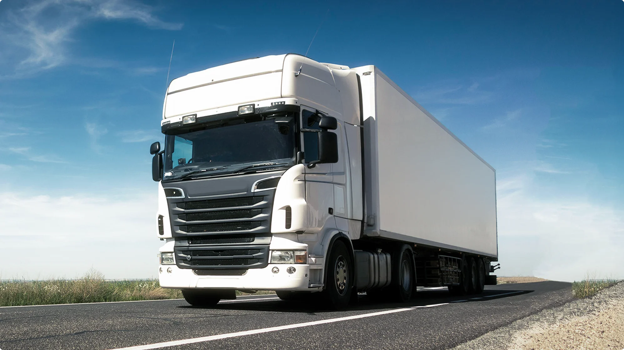 White articulated lorry driving on a rural road under clear blue sky, with fields visible on either side.
