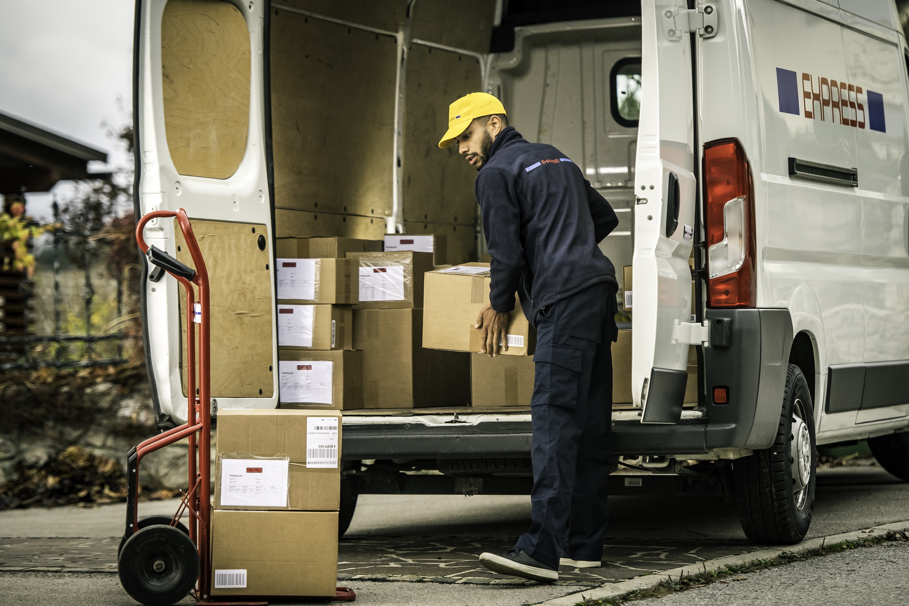 A delivery driver in a yellow cap unloads cardboard parcels from a white express van onto a red sack truck.