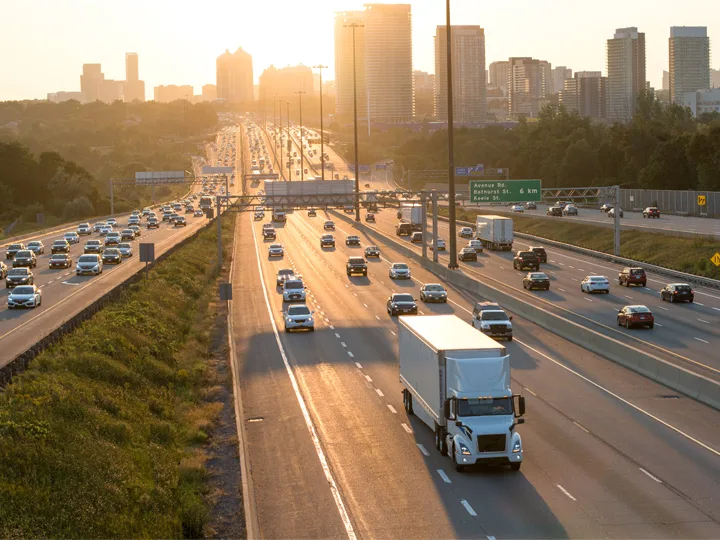 Busy highway with traffic flowing toward city skyline at sunset, white semi truck prominent in foreground